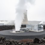 Steam rises from a geothermal well pad near a geothermal power plant, set among rugged volcanic rocks under an overcast sky.