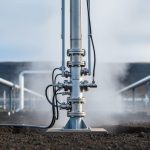 Steam rising from a geothermal wellhead and drilling equipment in a rugged field under soft daylight.